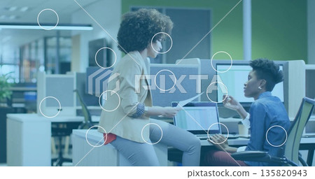 Two women in suits discussing printed reports while pointing at laptop charts in open-plan office 135820943