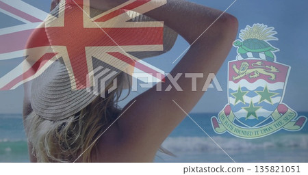 Swimwear-clad woman holding sunhat gazing at horizon on beach, with Union Jack and coat of arms 135821051