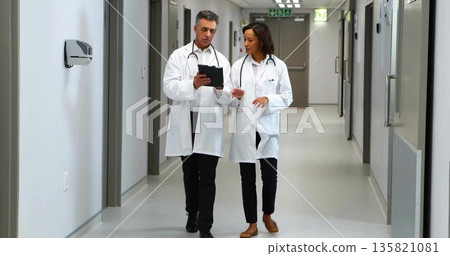 Walking doctor duo reviewing clipboard notes in hospital corridor, with stethoscopes and lab coats 135821081