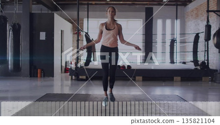 Jumping woman in pink top, leggings swinging jump rope on boxing gym mat, with boxing ring Jumping woman in pink top, leggings swinging jump rope on boxing gym mat, with boxing ring 135821104