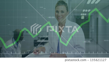 Smiling doctor wearing lab coat writing on clipboard in clinic corridor with green graph overlay Smiling doctor wearing lab coat writing on clipboard in clinic corridor with green graph overlay 135821127