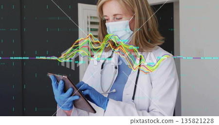 Reviewing tablet, doctor wearing coat, mask, stethoscope and gloves in exam room, monitoring graphs 135821228