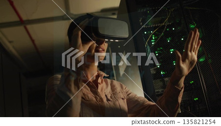 Interacting IT tech wearing shirt with VR headset in data center corridor, with blinking rack LEDs Interacting IT tech wearing shirt with VR headset in data center corridor, with blinking rack LEDs 135821254