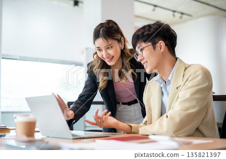 Smiling asian businesswoman looking at laptop talking with intern coworker at wooden table in office Smiling asian businesswoman looking at laptop talking with intern coworker at wooden table in office 135821397
