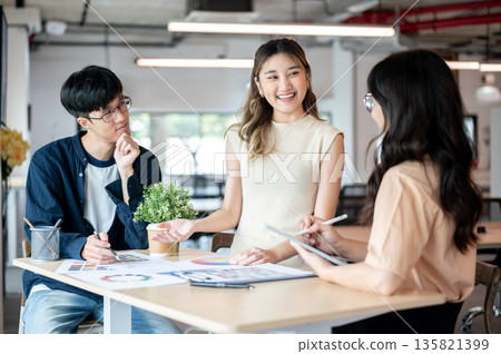 Smiling asian woman designer talking with coworkers about color chart at wooden table in office. Smiling asian woman designer talking with coworkers about color chart at wooden table in office. 135821399