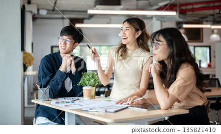 Asian woman designer holding pen smiling or laughing with coworkers at wooden table in the office. 135821410