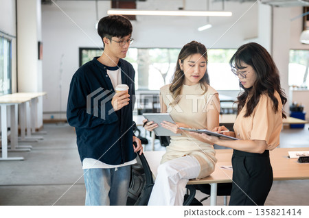 Asian woman holding tablet looking at clipboard in coworker's hand while sitting on table in office. Asian woman holding tablet looking at clipboard in coworker's hand while sitting on table in office. 135821414