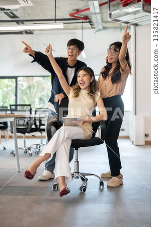 Asian woman holding tablet and raising a hand with coworkers while sitting on swivel chair in office 135821417