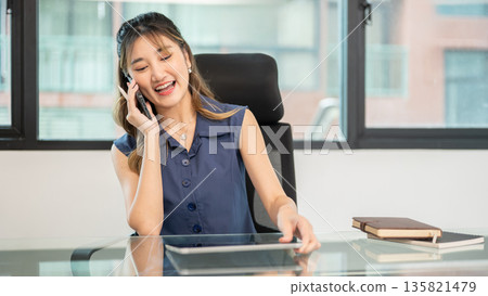Asian woman office worker putting down tablet as talking on phone while sit at glass table in office Asian woman office worker putting down tablet as talking on phone while sit at glass table in office 135821479