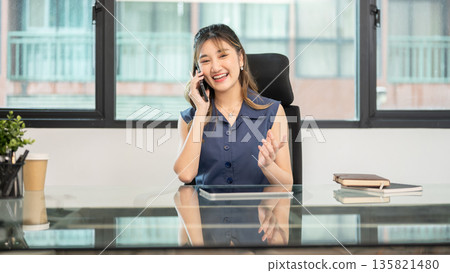 Asian woman office worker talking on phone aside tablet while sitting at glass table in the office. Asian woman office worker talking on phone aside tablet while sitting at glass table in the office. 135821480