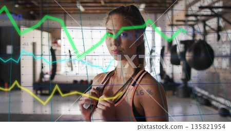 Training athlete in sportswear holding jump rope handles in gym, with punching bags, digital charts Training athlete in sportswear holding jump rope handles in gym, with punching bags, digital charts 135821954