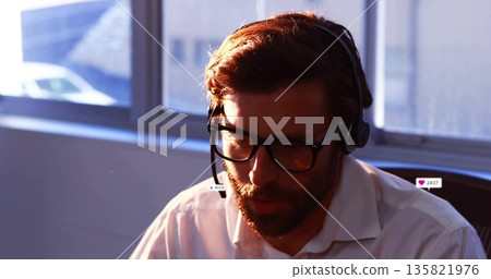 Bearded man wearing headset and eyeglasses reading customer message at office desk near window 135821976
