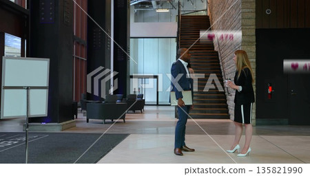 Standing colleagues wearing suits holding coffee cup and laptop in office lobby with signboard 135821990