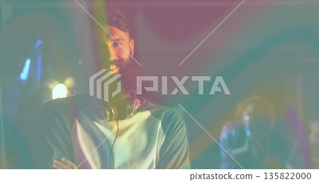 Smiling bearded DJ crossing arms on studio stage, with headphones, microphone and colored lights Smiling bearded DJ crossing arms on studio stage, with headphones, microphone and colored lights 135822000