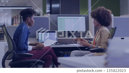 Holding reports and coffee cup, two colleagues reviewing spreadsheet data in open-plan office 135822023