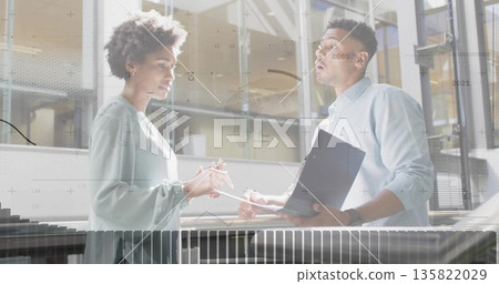 Discussing data two colleagues reviewing reports in modern atrium, with folder pen railing glass 135822029
