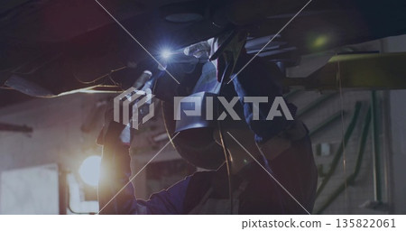 Welding welder wearing helmet guiding torch beneath raised vehicle chassis in workshop, with lamp Welding welder wearing helmet guiding torch beneath raised vehicle chassis in workshop, with lamp 135822061
