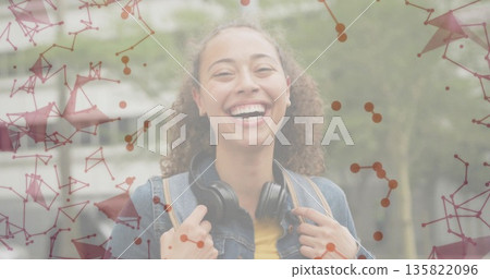 Smiling woman holding backpack walking on path in denim jacket, headphones under network overlay 135822096