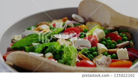 White ceramic bowl resting on light tabletop, with salad, tomatoes, croutons, wooden tongs 135822102