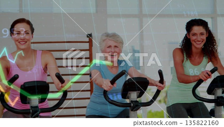 Cycling trio of women riding exercise bikes in studio, with bike displays showing performance data 135822160