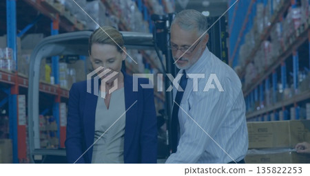 Colleagues wearing business attire checking boxes in warehouse aisle on shelving, with forklift 135822253