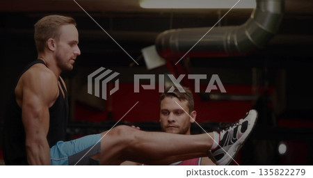 Spotter supporting man wearing black tank top doing straight-leg raise at gym area, with dip bars 135822279