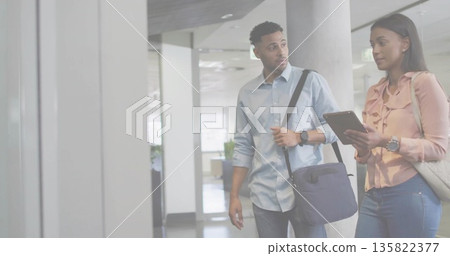 Walking two business colleagues reviewing tablet computer in office corridor, with messenger bag Walking two business colleagues reviewing tablet computer in office corridor, with messenger bag 135822377
