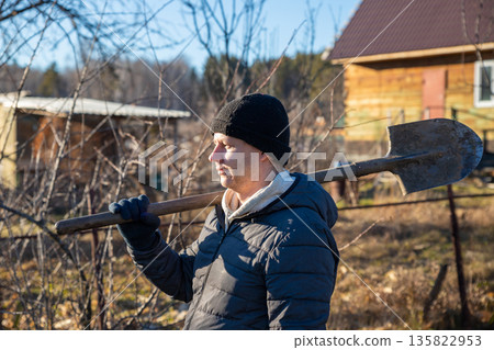Man carrying a shovel on his shoulder to work at a community garden, concept of labor, rehabilitation, and correctional work. Rural spring or autumn outdoor activity. Man carrying a shovel on his shoulder to work at a community garden, concept of labor, rehabilitation, and correctional work. Rural spring or autumn outdoor activity. 135822953