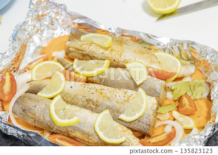 Prerape raw spiced hake for a bake in the oven with lemon, tomatoes, carrots, and onions in foil. Delicious fish for lunch or dinner, perfect for a diet. Tasty and fresh  hake fish for bake with 135823123