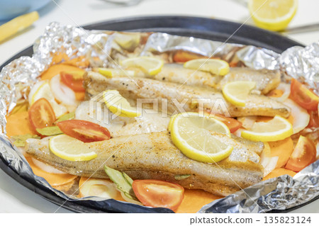 Prerape raw spiced hake for a bake in the oven with lemon, tomatoes, carrots, and onions in foil. Delicious fish for lunch or dinner, perfect for a diet. Tasty and fresh  hake fish for bake with 135823124