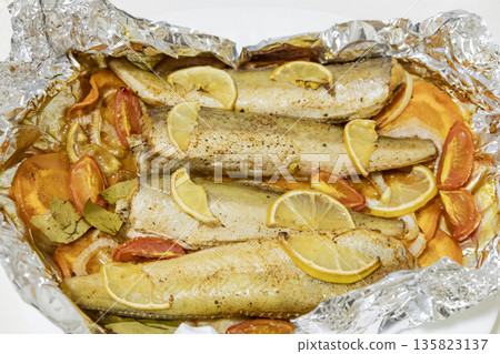 Spiced hake baked in the oven with lemon, tomatoes, carrots, and onions in foil. Delicious fish for lunch or dinner, perfect for a diet. Tasty hake fish baked with vegetables. 135823137