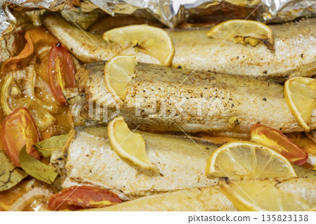 Spiced hake baked in the oven with lemon, tomatoes, carrots, and onions in foil. Delicious fish for lunch or dinner, perfect for a diet. Tasty hake fish baked with vegetables. 135823138