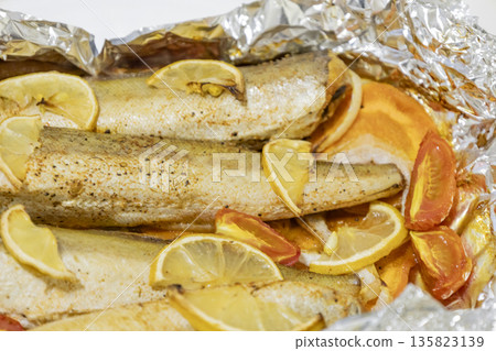 Spiced hake baked in the oven with lemon, tomatoes, carrots, and onions in foil. Delicious fish for lunch or dinner, perfect for a diet. Tasty hake fish baked with vegetables. 135823139