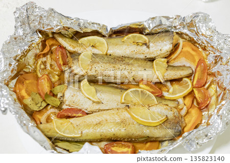Spiced hake baked in the oven with lemon, tomatoes, carrots, and onions in foil. Delicious fish for lunch or dinner, perfect for a diet. Tasty hake fish baked with vegetables. 135823140