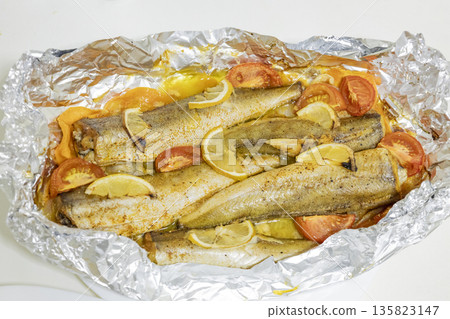 Spiced hake baked in the oven with lemon, tomatoes, carrots, and onions in foil. Delicious fish for lunch or dinner, perfect for a diet. Tasty hake fish baked with vegetables. 135823147