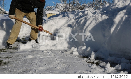 A man, visible without a face, clears large snowdrifts in front of a house with a wooden shovel in sunny weather. Manual snow removal from a house's surrounding area after a storm. 135823662