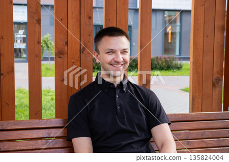 Caucasian man smiling happily while sitting on a wooden bench outdoors. Relaxed mood for urban lifestyle and leisure concept. Caucasian man smiling happily while sitting on a wooden bench outdoors. Relaxed mood for urban lifestyle and leisure concept. 135824004