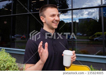 Young Caucasian man holds paper cup of coffee and waves his hand, smiling happily on city street. Businessman or office worker enjoying coffee break. Date, lifestyle and communication. Young Caucasian man holds paper cup of coffee and waves his hand, smiling happily on city street. Businessman or office worker enjoying coffee break. Date, lifestyle and communication. 135824005