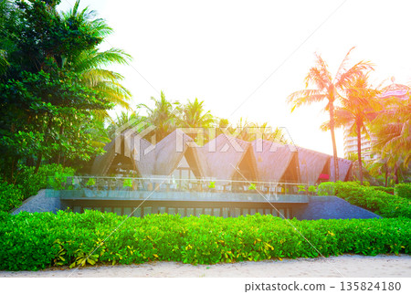Beachfront restaurant with thatched roofs with palm trees at sunset.  135824180