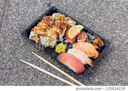 A delicious sushi platter features assorted rolls, sashimi, and wasabi, all presented on a black tray. The meal is set on a gray stone surface, reflecting a casual dining experience 135824219