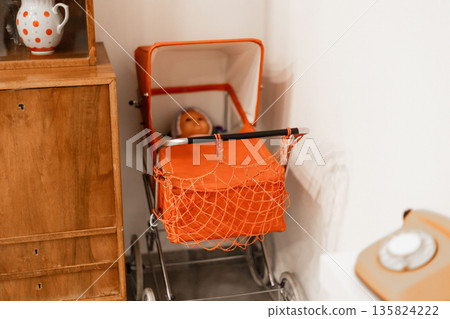 A charming vintage doll stroller, featuring a bright orange color, holds a doll inside. The setting reflects a cozy and nostalgic atmosphere, with wooden furniture and decor 135824222
