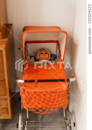 A bright orange baby stroller holds a doll dressed in a bonnet. It is positioned next to wooden furniture in a cozy indoor space, illuminated by natural light 135824223