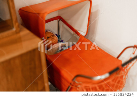 A vintage doll lies quietly in a bright orange stroller, surrounded by a warm and inviting room setting, highlighting a nostalgic atmosphere A vintage doll lies quietly in a bright orange stroller, surrounded by a warm and inviting room setting, highlighting a nostalgic atmosphere 135824224
