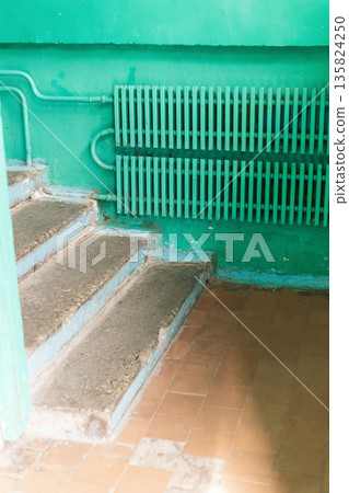 The interior of an old house reveals worn-out steps leading to a bright green wall. A rusty radiator adds to the neglected atmosphere. Dust and marks indicate years of disrepair The interior of an old house reveals worn-out steps leading to a bright green wall. A rusty radiator adds to the neglected atmosphere. Dust and marks indicate years of disrepair 135824250