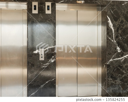 Two elevator doors stand in a building with dark marble walls. The setting shows a clean and modern environment for accessing different floors of the structure Two elevator doors stand in a building with dark marble walls. The setting shows a clean and modern environment for accessing different floors of the structure 135824255