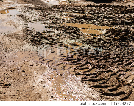 Wet mud covers the ground at a construction site. Tire tracks show recent vehicle movement. Water collects in puddles, reflecting light. The scene captures the aftermath of rain Wet mud covers the ground at a construction site. Tire tracks show recent vehicle movement. Water collects in puddles, reflecting light. The scene captures the aftermath of rain 135824257
