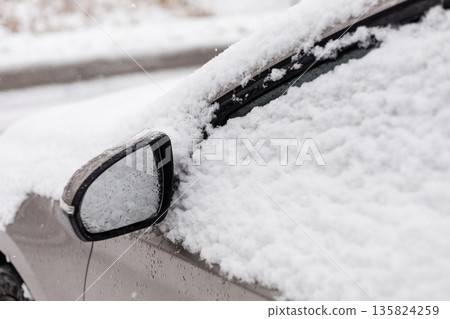 A parked car is covered with a thick layer of fresh snow. Large snowflakes fall gently, blanketing the vehicle and surrounding area. The scene shows a quiet street in winter 135824259
