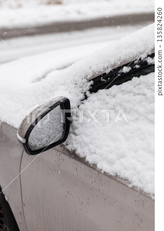 A car is parked on a street surrounded by snow. The snow has accumulated on the roof and side mirror. It is winter, and the ground is covered in white snow 135824260