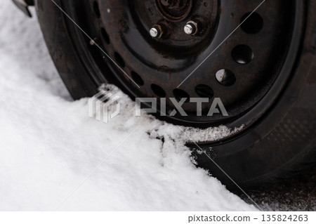 A car tire is partly covered in snow on a city street during winter. Snow blankets the ground, showing a scene of cold weather and difficult driving conditions A car tire is partly covered in snow on a city street during winter. Snow blankets the ground, showing a scene of cold weather and difficult driving conditions 135824263