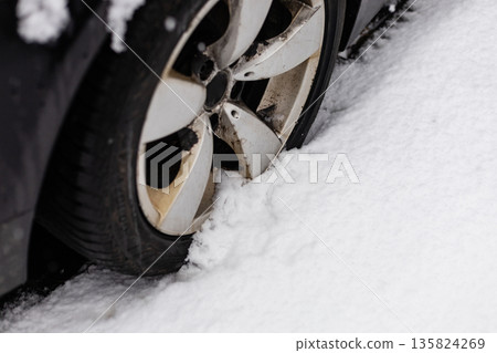 A car tire is partially buried in snow, showing signs of struggle. The vehicle is stuck on a snowy street. Snow covers the ground and parts of the tire A car tire is partially buried in snow, showing signs of struggle. The vehicle is stuck on a snowy street. Snow covers the ground and parts of the tire 135824269